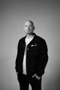 Portrait of Pony Lawson, tattoo artist at Inked Tattoo Studios, wearing a black jacket and standing confidently against a neutral background, reflecting the artistry and personal journey shared in the interview.