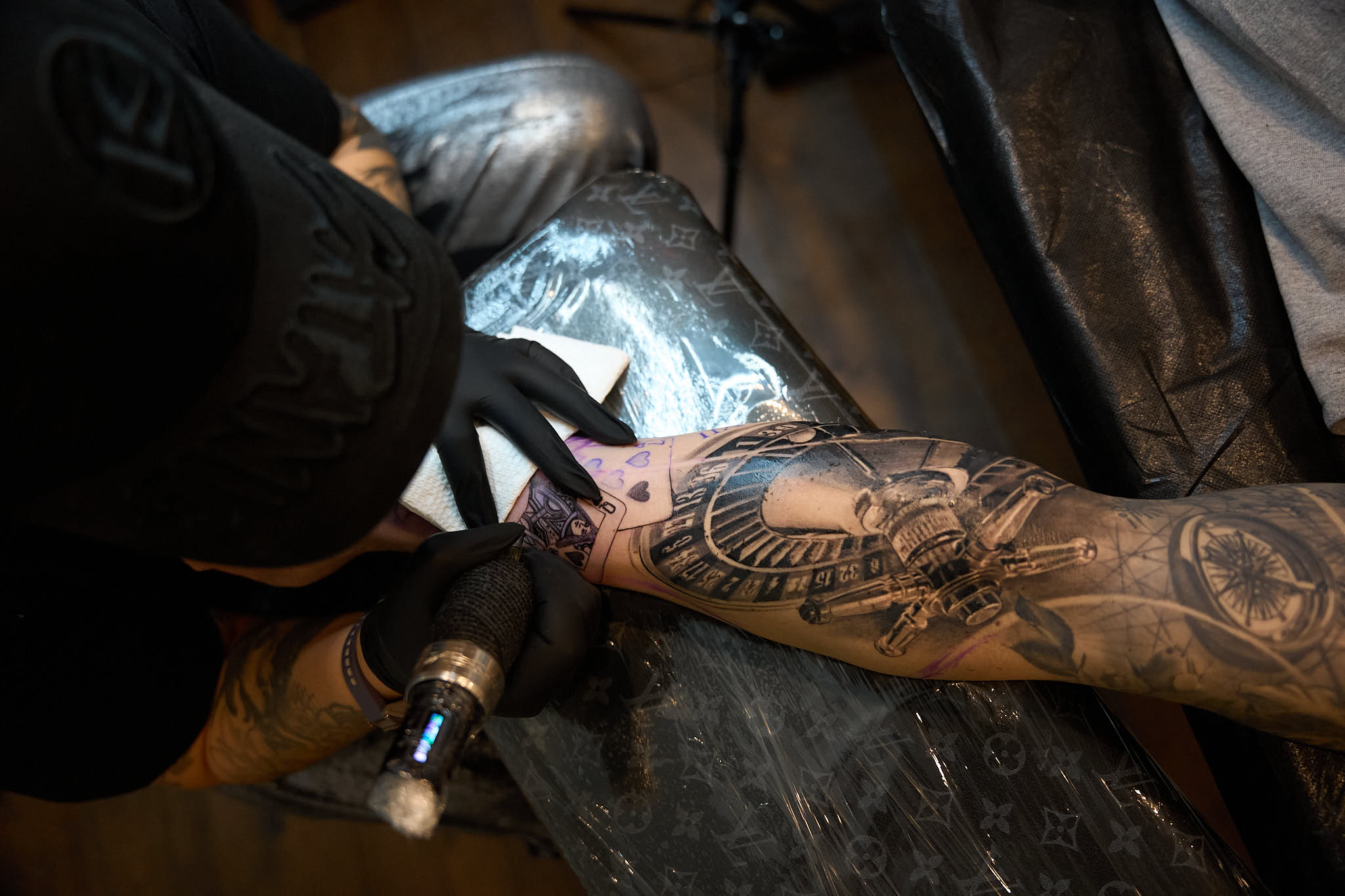 Tattoo artist working on a detailed sleeve tattoo design featuring intricate elements, including a roulette wheel and compass, in a professional studio setting at Inked Tattoo Studios.