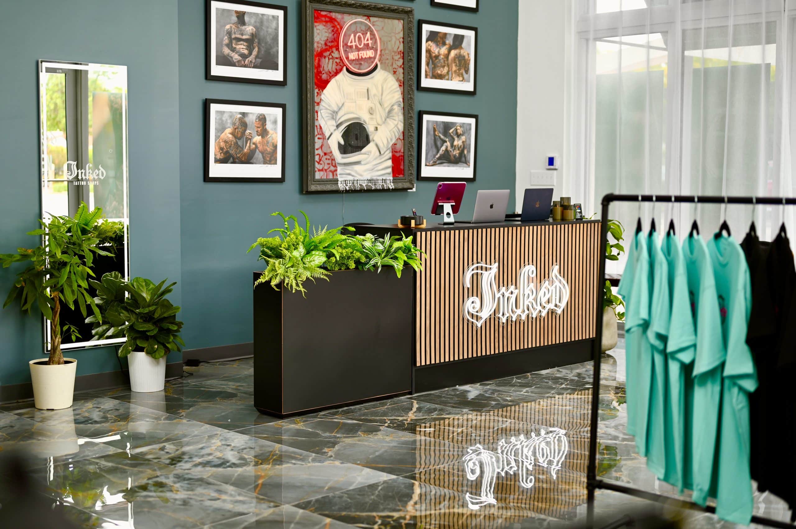 Inked Tattoo Studios reception area featuring a stylish counter with a neon "Inked" sign, surrounded by framed tattoo art, plants, and merchandise display.