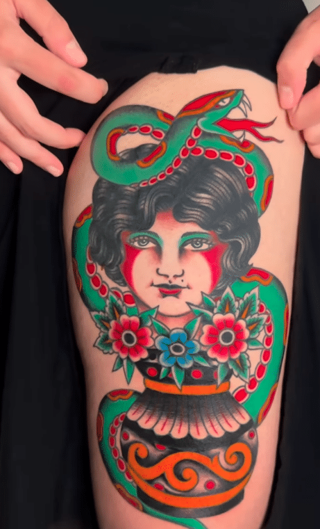 A large thigh tattoo in American traditional style of pin-up girl head coming out of a vase of flowers and surrounded by a snake