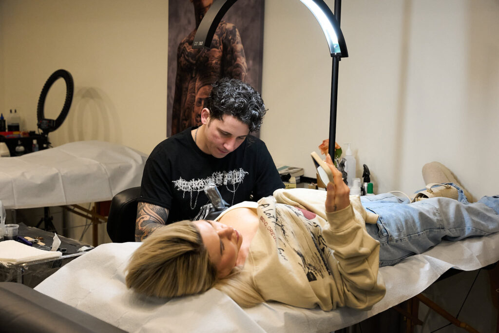 Woman lying down and looking at her phone while getting a tattoo 