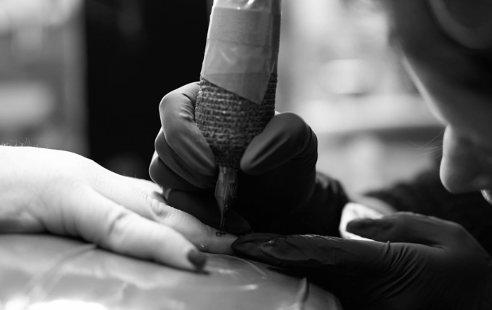 Black and white photo of a tattoo artist tattooing a small design on a client’s finger.