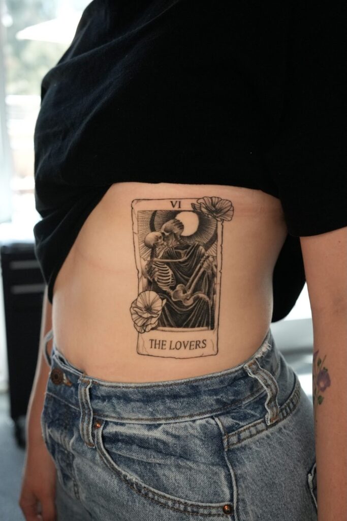 Black ink rib tattoo of The Lovers tarot card, showing two skeletons embracing with flowers and a moon above.