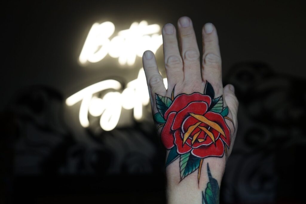 Bright red rose tattoo on the back of the hand in traditional style with heavy black outlines and green leaves.