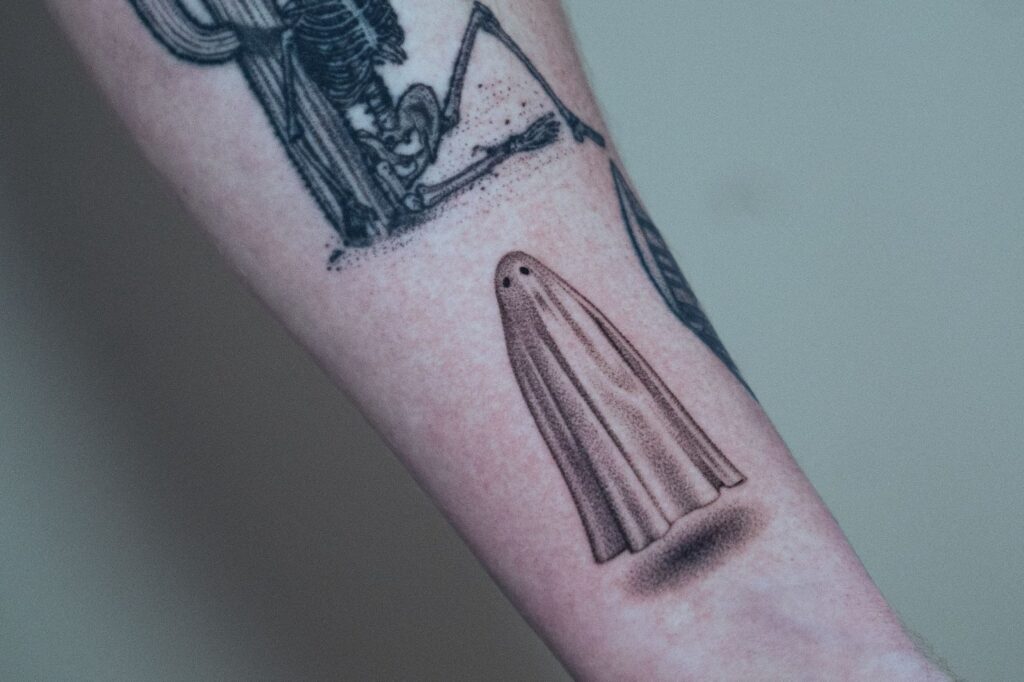 Minimalist black and grey tattoo of a small sheet-draped ghost, floating above the skin with a soft shadow beneath.
