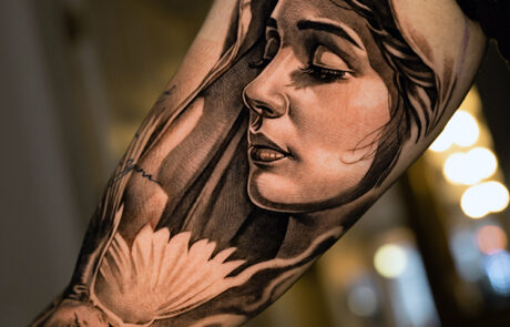 Black and grey tattoo on a person’s upper arm featuring a detailed portrait of a woman with flowing hair and a dove in flight below her face.