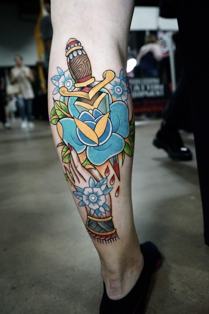 Colorful traditional-style tattoo on the back of a person’s calf, depicting a hand holding a dagger surrounded by blue flowers and leaves with drops of red ink below.