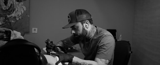 A focused tattoo artist wearing a hat and gloves while working on a client’s arm in a studio setting.