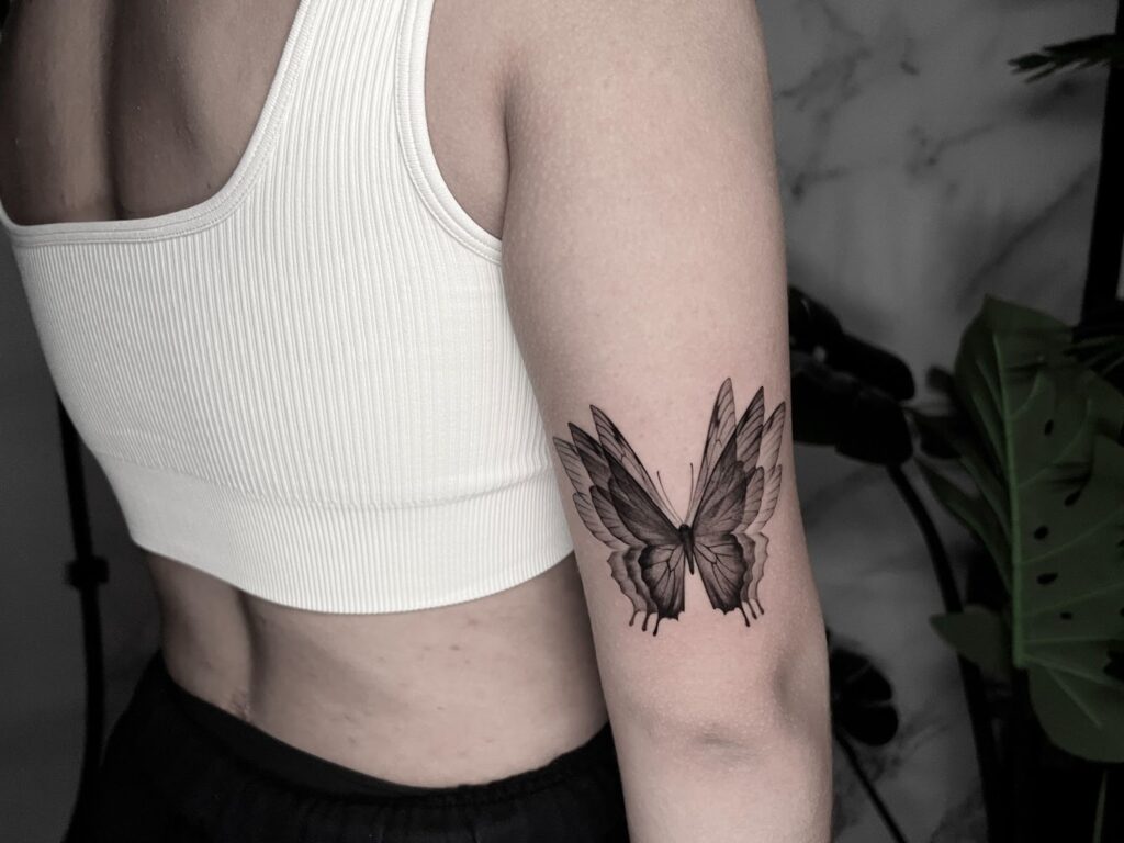 Black and grey butterfly tattoo on the back of a womanโs arm, featuring fine linework and delicate shading.