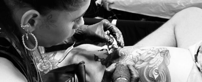 Black and white photo of a tattoo artist working on a client’s thigh at a busy tattoo convention, surrounded by other artists and attendees in the background.
