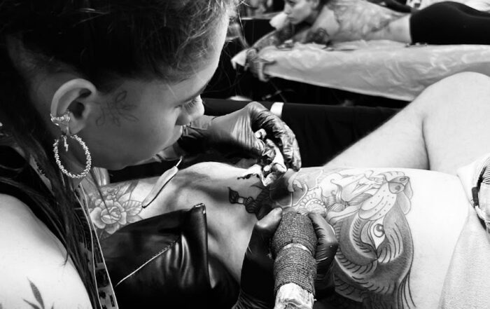 Black and white photo of a tattoo artist working on a client’s thigh at a busy tattoo convention, surrounded by other artists and attendees in the background.