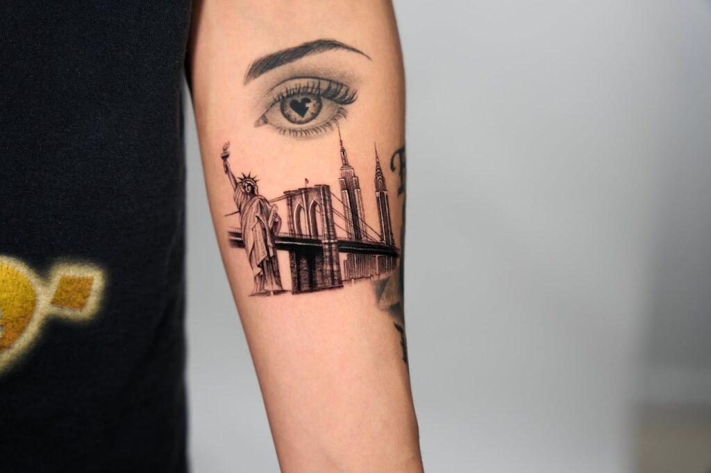 Detailed black and grey tattoo of the New York City skyline,ย including the Statue of Liberty, Empire State Building, and Brooklyn Bridge,ย on a personโs inner arm.