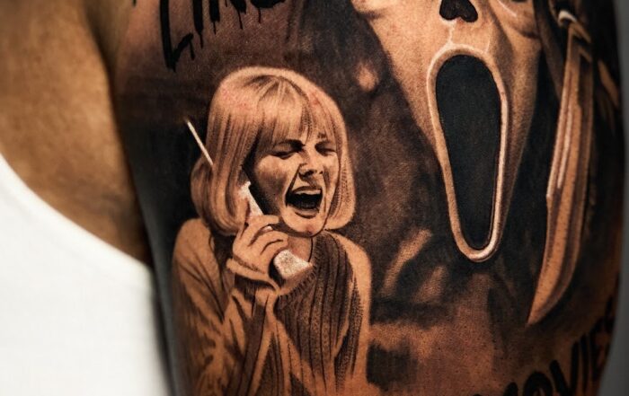 Black and grey upper-arm tattoo featuring the Ghostface mask from the movie “Scream” beside a woman holding a phone and screaming, with the text “Do you like scary movies” in dripping horror-style font.
