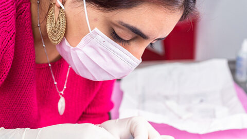 A piercer wearing gloves and a face mask performing a facial piercing on a person who is lying down with a protective drape over their eyes.