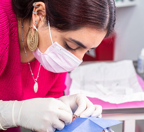 A piercer wearing gloves and a face mask performing a facial piercing on a person who is lying down with a protective drape over their eyes.