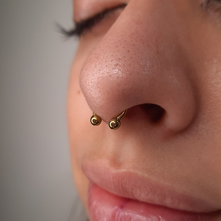 Close-up of a nose with a septum piercing wearing a circular gold ring with small gold ball ends.