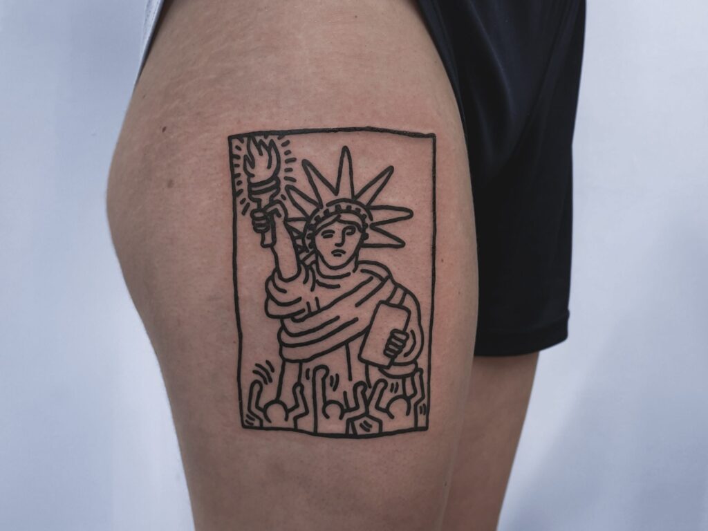 A Keith Haring-inspired linework tattoo of the Statue of Liberty on the thigh, drawn in ignorant style.
