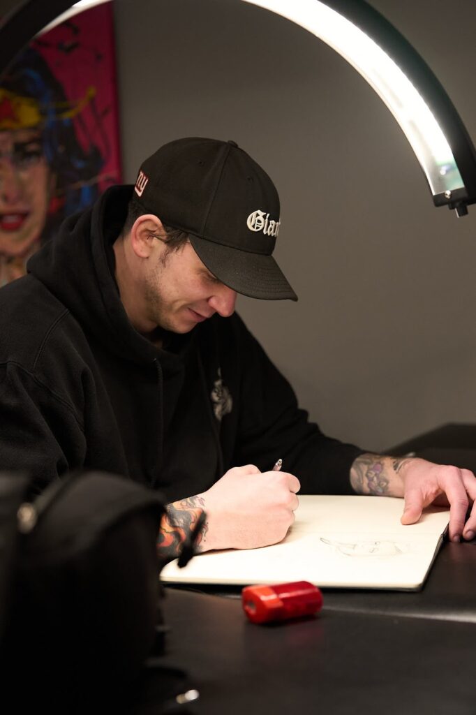 A tattoo artist sketching a design in a notebook under a bright studio lamp.