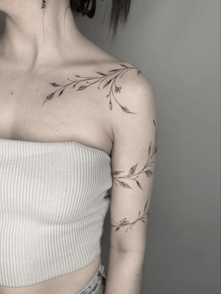 Delicate fine-line botanical tattoo wrapping across the shoulder and upper arm, featuring thin branches, leaves, and small buds.