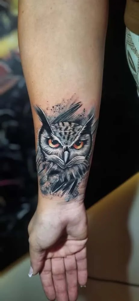 Black and grey realism tattoo of an owl with vivid orange eyes, detailed feathers, and textured shading on the forearm.
