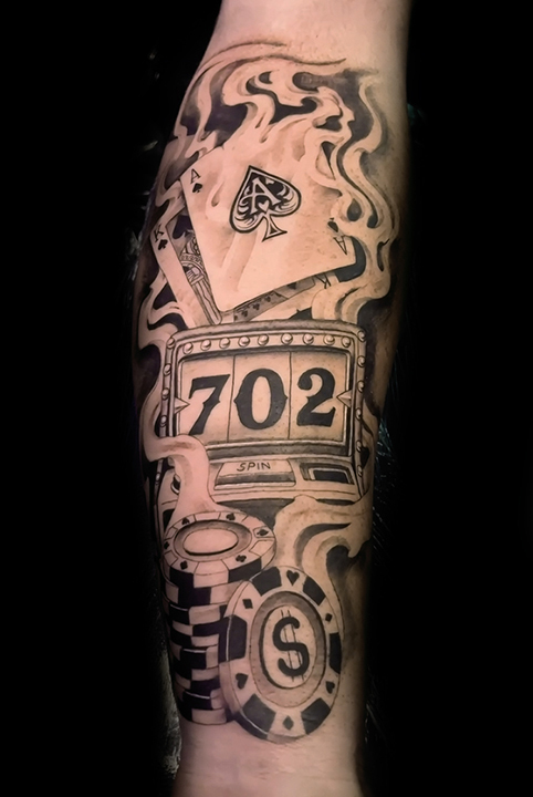 Black and grey tattoo featuring playing cards, a slot machine, and casino chips, with the number "702" and smoke accents on the forearm.