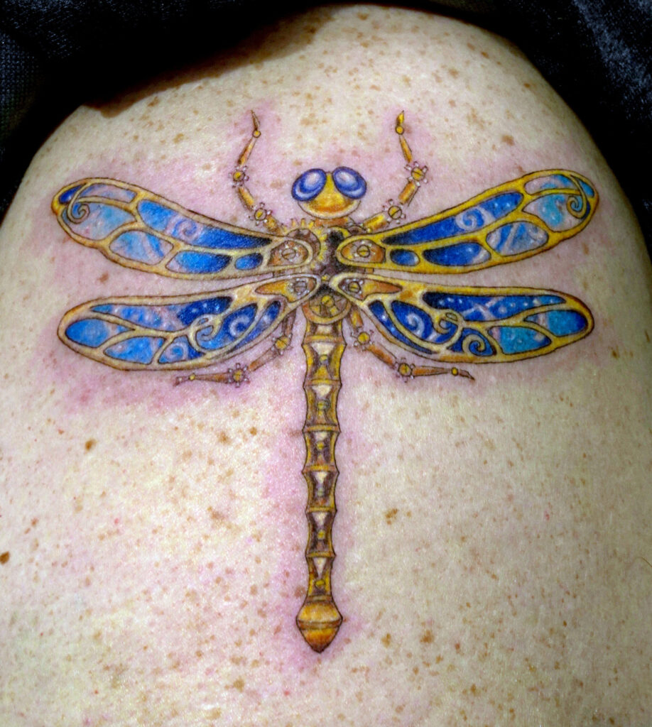 Close-up of a detailed dragonfly tattoo with blue and gold wings on freckled skin, showing fine linework and shading.