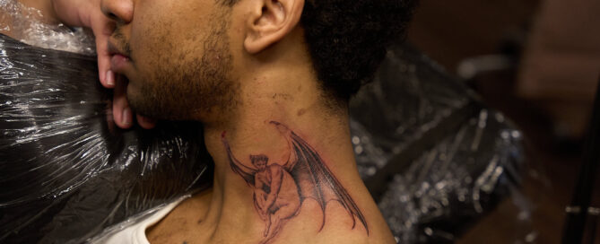 Side profile of a person with a fine-line winged figure tattoo on the side of the neck, photographed in an Inked tattoo studio.