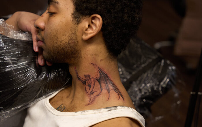 Side profile of a person with a fine-line winged figure tattoo on the side of the neck, photographed in an Inked tattoo studio.
