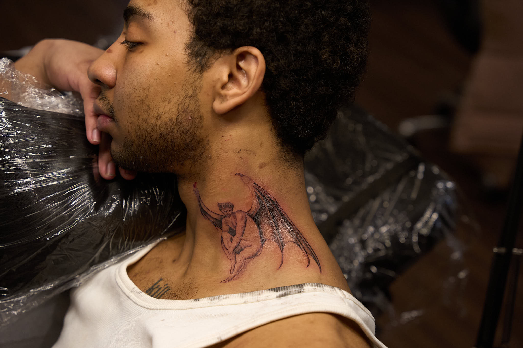 Side profile of a person with a fine-line winged figure tattoo on the side of the neck, photographed in an Inked tattoo studio.