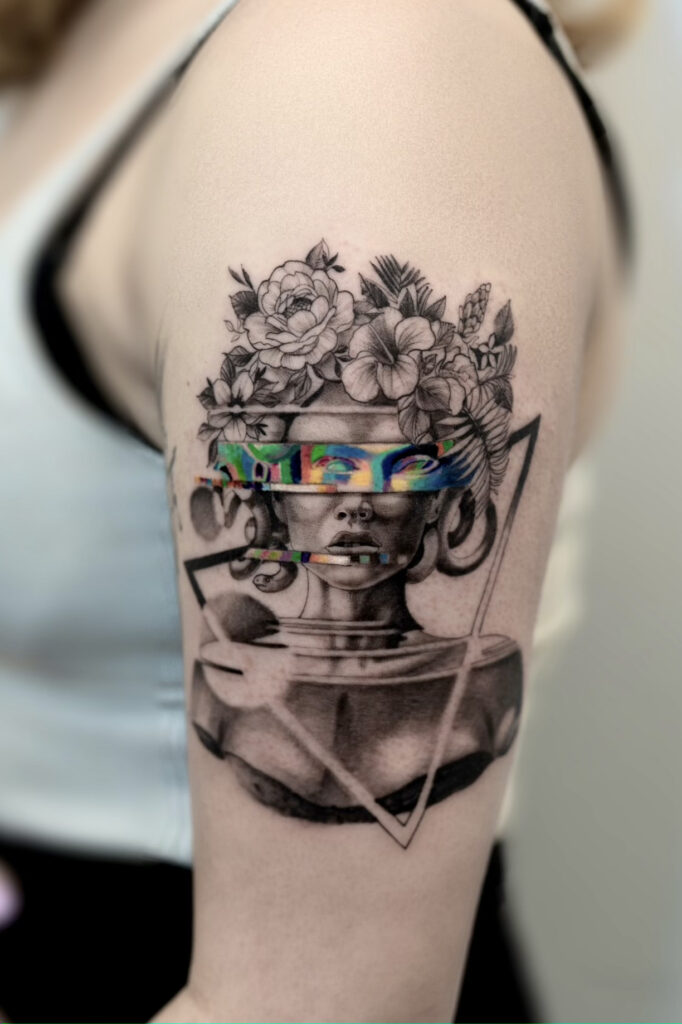 Black and gray upper arm tattoo featuring a surreal female bust with floral elements, geometric linework, and a colorful glitch-style band across the eyes.