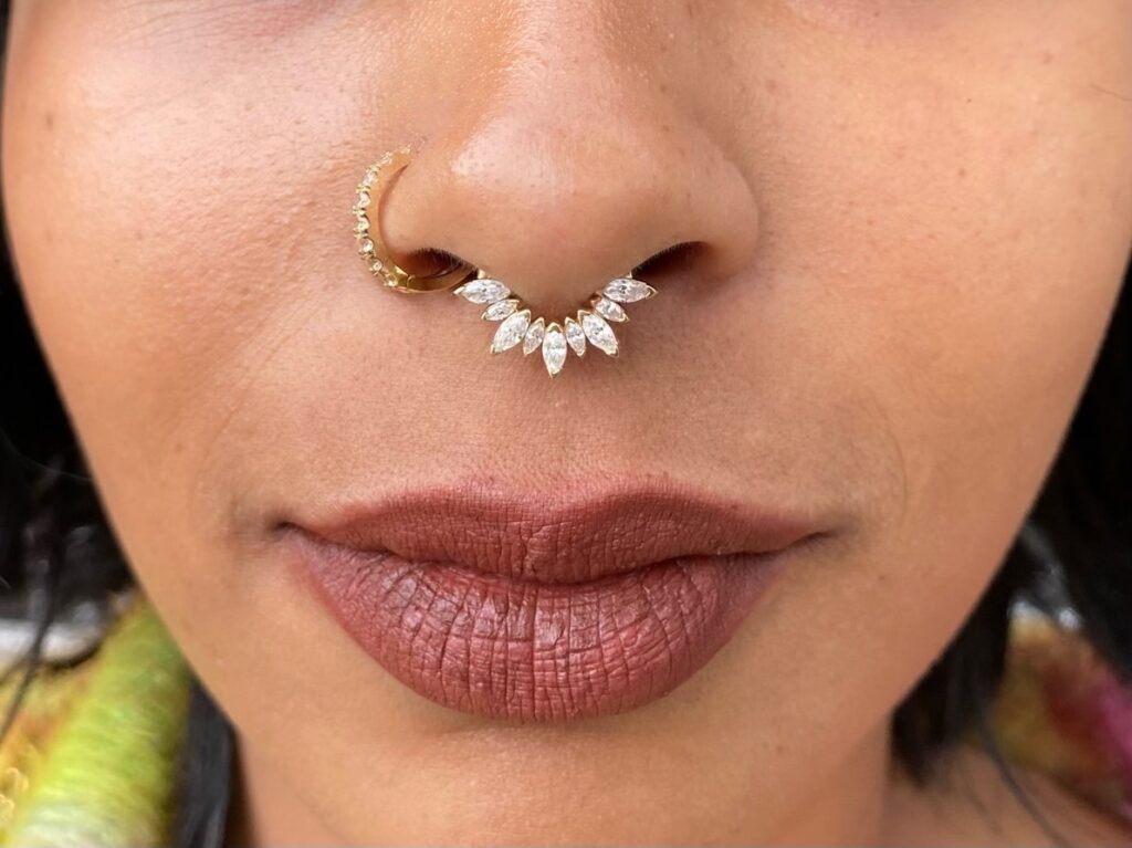 Close-up of a personโs nose and lips featuring a gold septum ring with marquise-shaped clear gemstones arranged in a curved fan design.