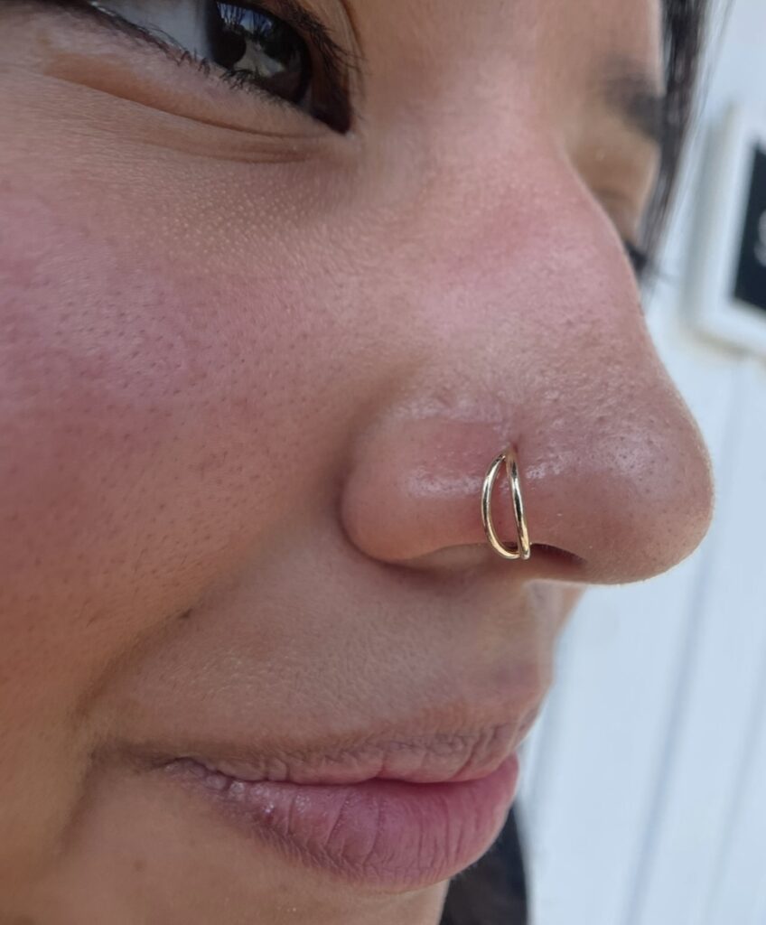 Side profile close-up of a nose with a thin gold double-hoop nostril ring.