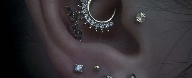 Close-up of an ear with multiple gold piercings, including a crescent-shaped hoop with small clear gemstones, several stud piercings, and a gold hoop in the lobe.