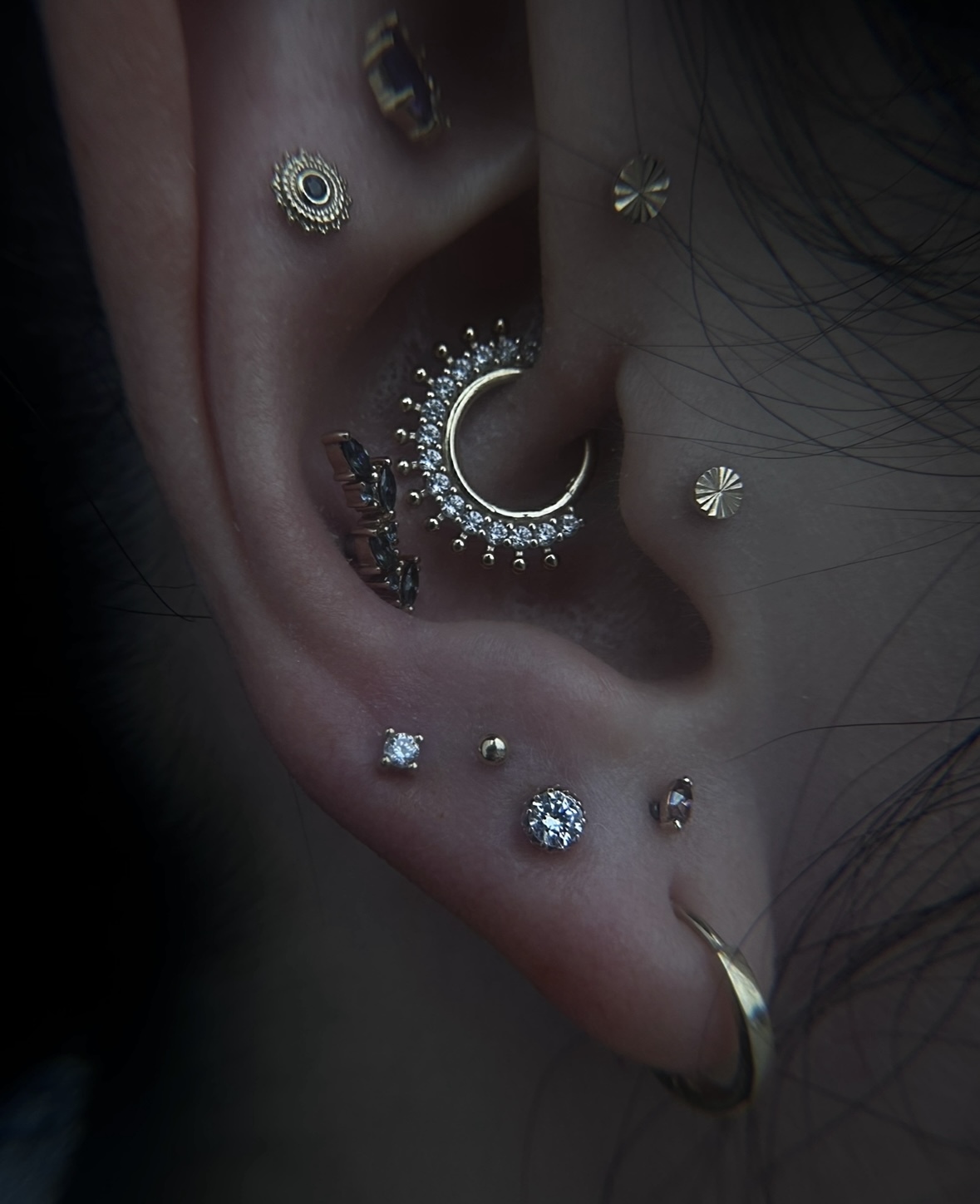 Close-up of an ear with multiple gold piercings, including a crescent-shaped hoop with small clear gemstones, several stud piercings, and a gold hoop in the lobe.