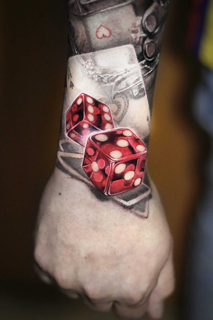 Realistic tattoo of red dice with white dots, placed near playing cards with a soft shaded background on the wrist.