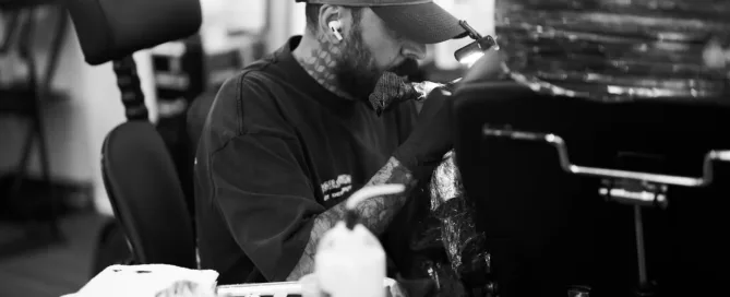 A black-and-white close-up of a focused tattoo artist wearing a baseball cap and wireless earbuds while working on a client.
