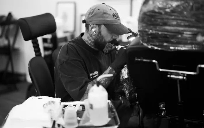 A black-and-white close-up of a focused tattoo artist wearing a baseball cap and wireless earbuds while working on a client.