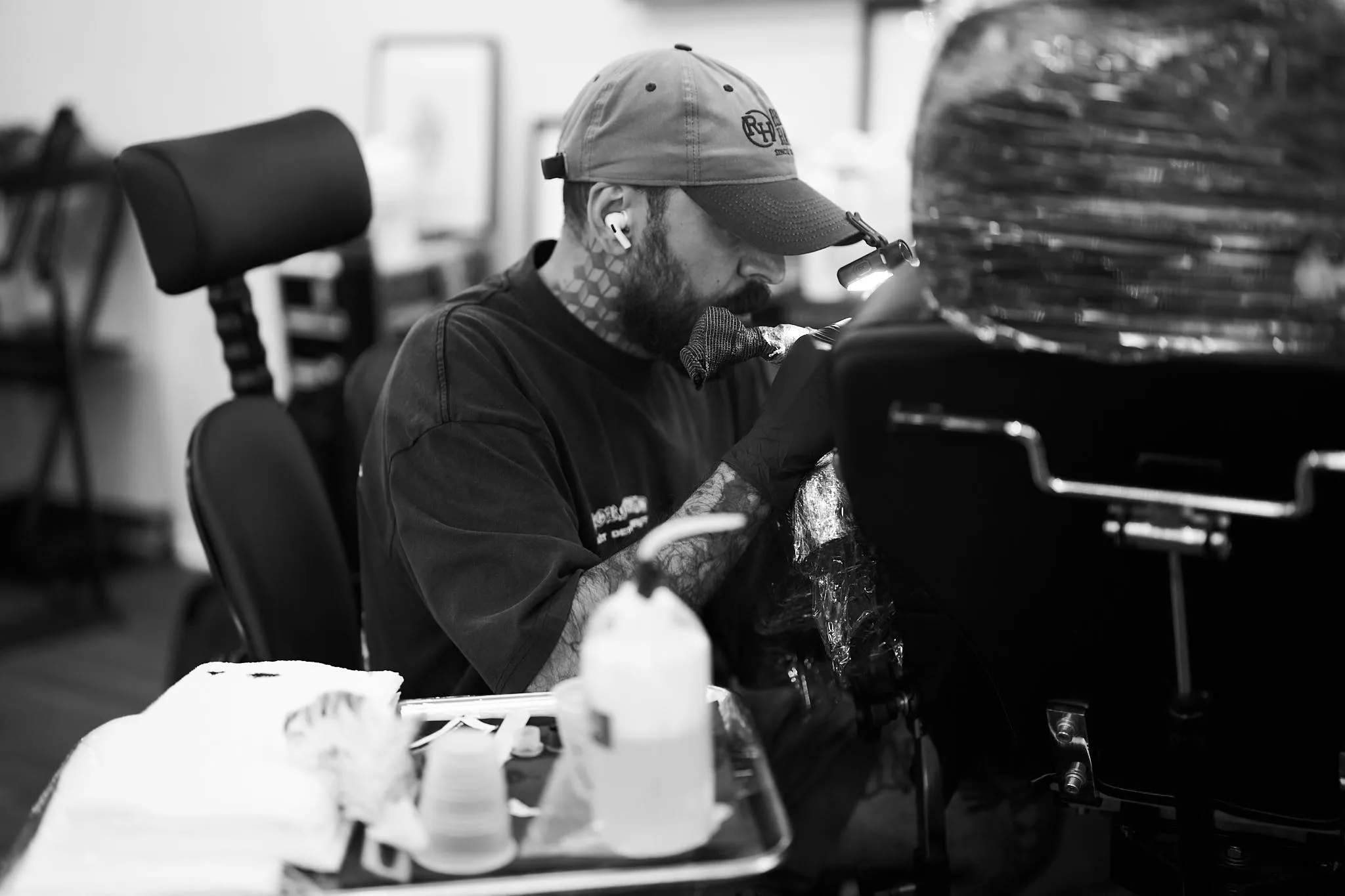 1c759006-e2bf-4e19-9348-15e78b8089f5 A black-and-white close-up of a focused tattoo artist wearing a baseball cap and wireless earbuds while working on a client.