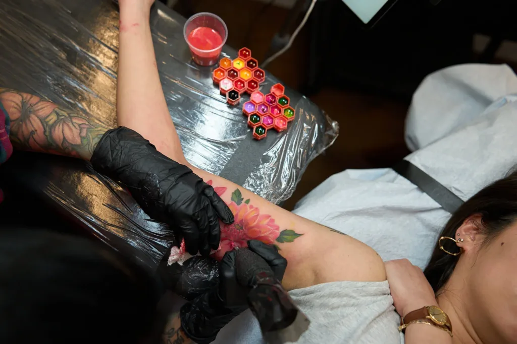 An overhead view of a gloved artist tattooing a vibrant pink floral design on a client's inner arm next to a palette of colorful inks.