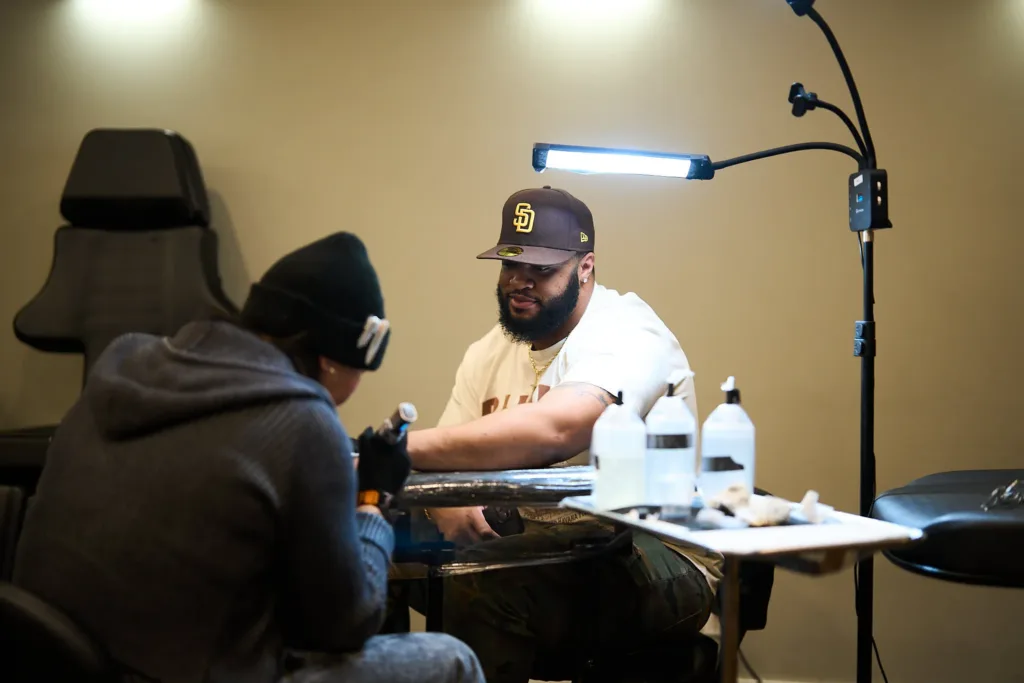 A tattoo artist in a black beanie works on a client’s forearm under a bright task light in a studio.