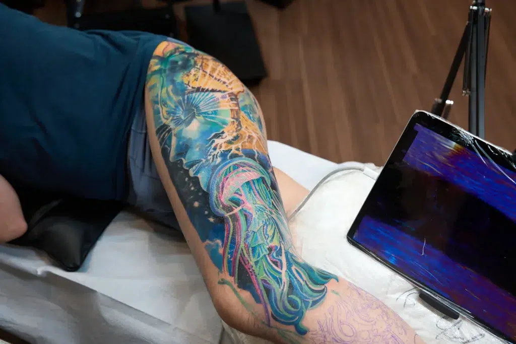 High-angle shot of a large, vibrant, blue and multicolored surrealist tattoo on a person's thigh, depicting a face and a jellyfish.