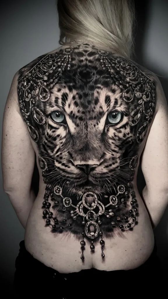 An intricate black-and-grey back tattoo of a leopard's face surrounded by ornate, hanging jewelry and beadwork.