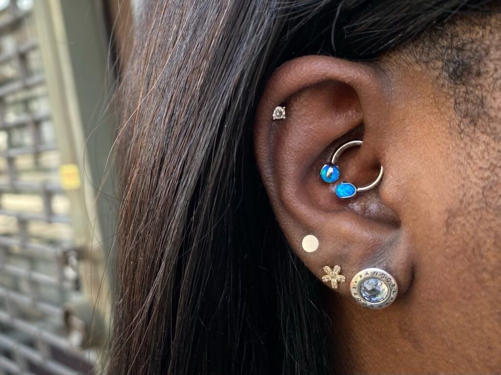 lose-up of an ear with multiple piercings, including a blue gemstone ring, small gold studs, and a cartilage piercing.