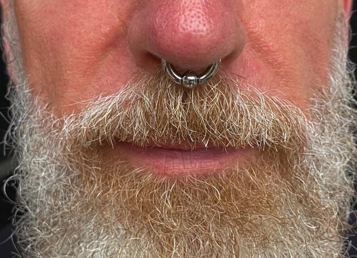 Close-up of a septum piercing with a silver horseshoe ring worn beneath the nose, framed by facial hair.