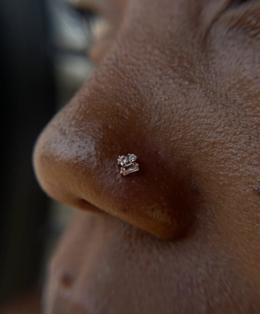 Close-up side view of a nostril piercing featuring a small cluster of clear gemstones in a gold setting.