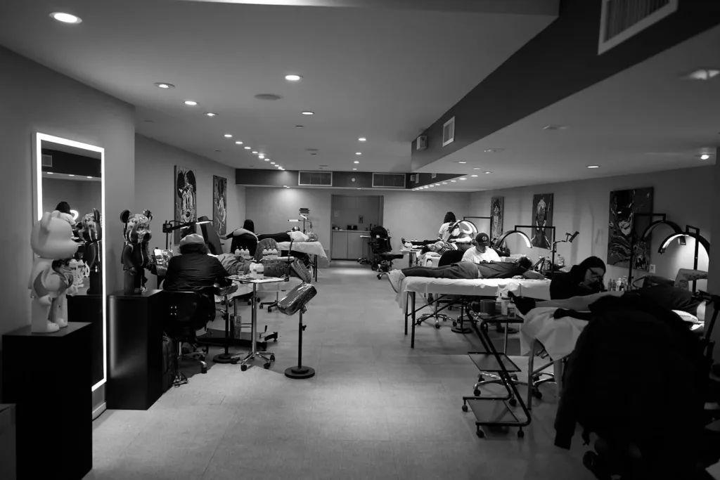 A wide, black-and-white view of a spacious tattoo studio with multiple artists working on clients at separate stations