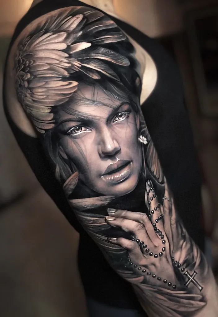 A stunning black-and-grey realism tattoo on a shoulder and upper arm, portraying a woman’s face framed by delicate feathers and a hand holding a rosary.