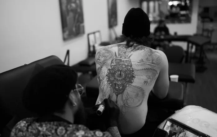 Black and white, wide shot of a tattoo artist working on a client&rsquo;s large, intricate back tattoo featuring a skull, mandala, and dragons.