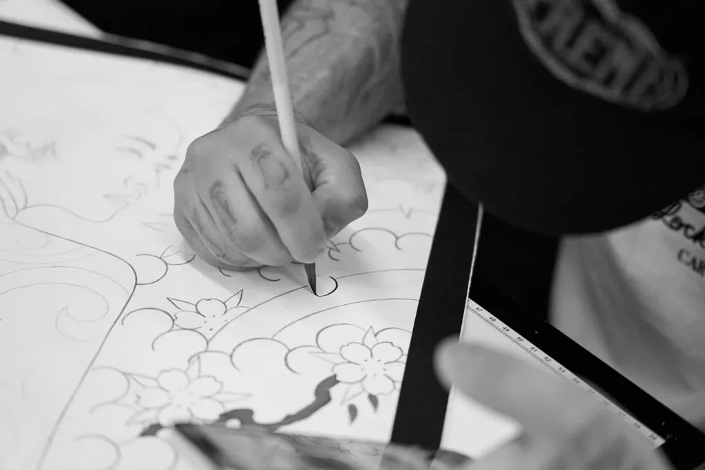 Black and white, close-up shot of an artist's hand using a thin brush to paint traditional floral and wave patterns onto a large canvas.