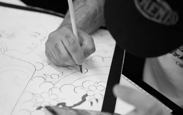 Black and white, close-up shot of an artist's hand using a thin brush to paint traditional floral and wave patterns onto a large canvas.